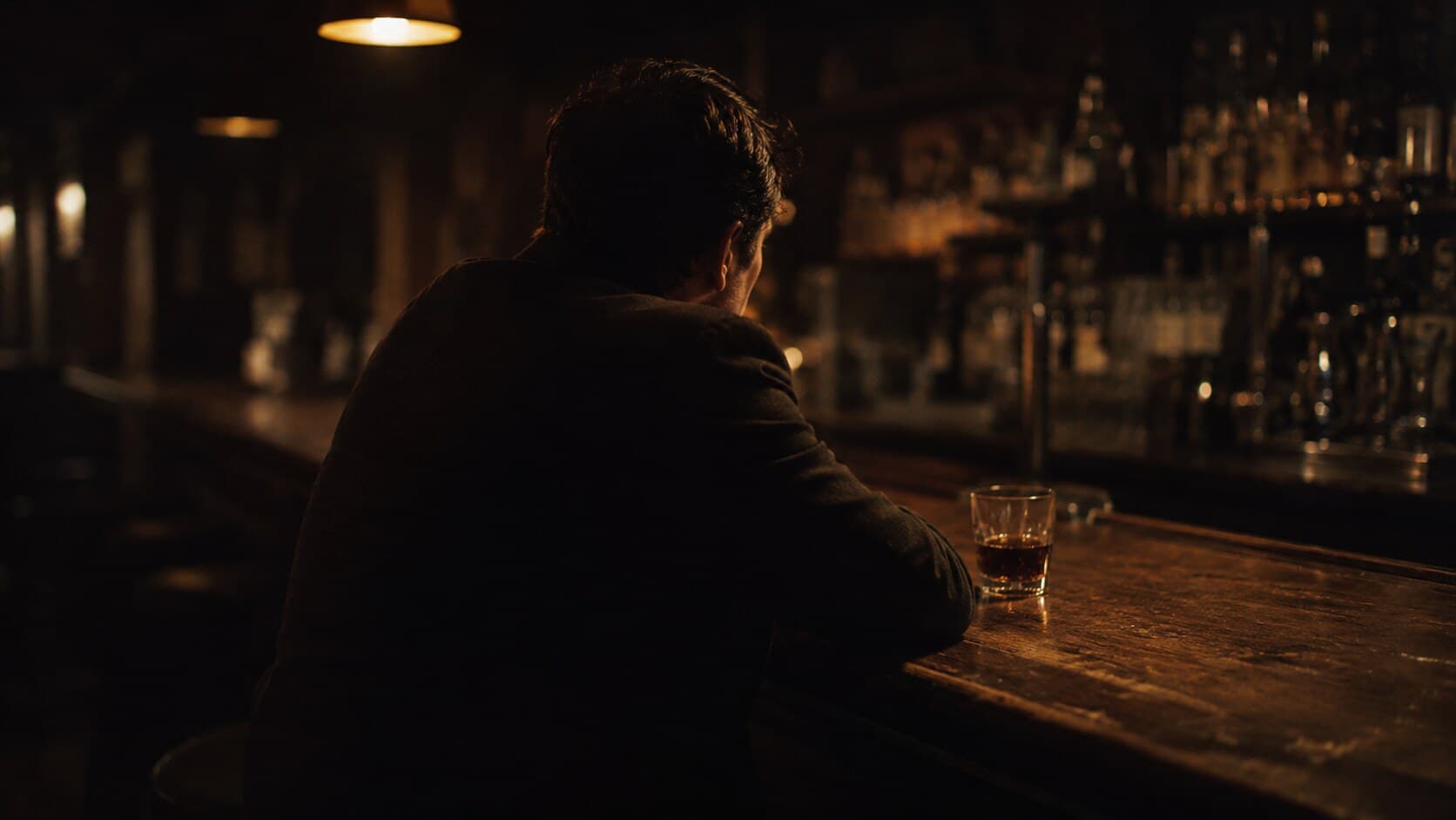 A man sits alone at a dimly lit bar, three-quarter rear profile, glass of whiskey on the counter.